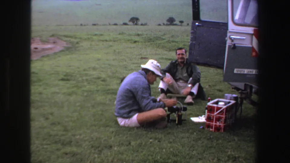A crouching man adjusts camera gear near a Toyota Land Cruiser while another man sits watching in the grassy South African landscape.