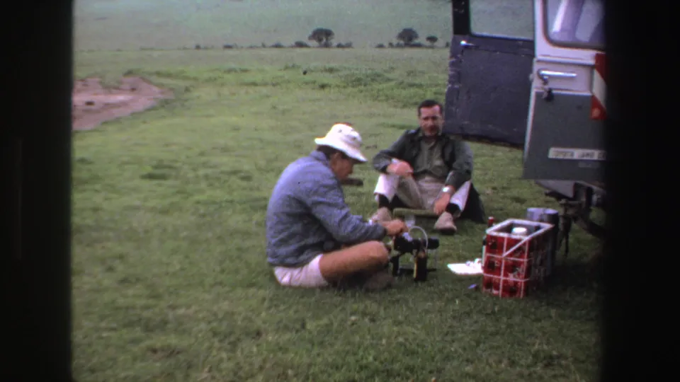 Two men remain beside the vehicle in a grassy South African field, with camera equipment and supplies arranged on the ground.