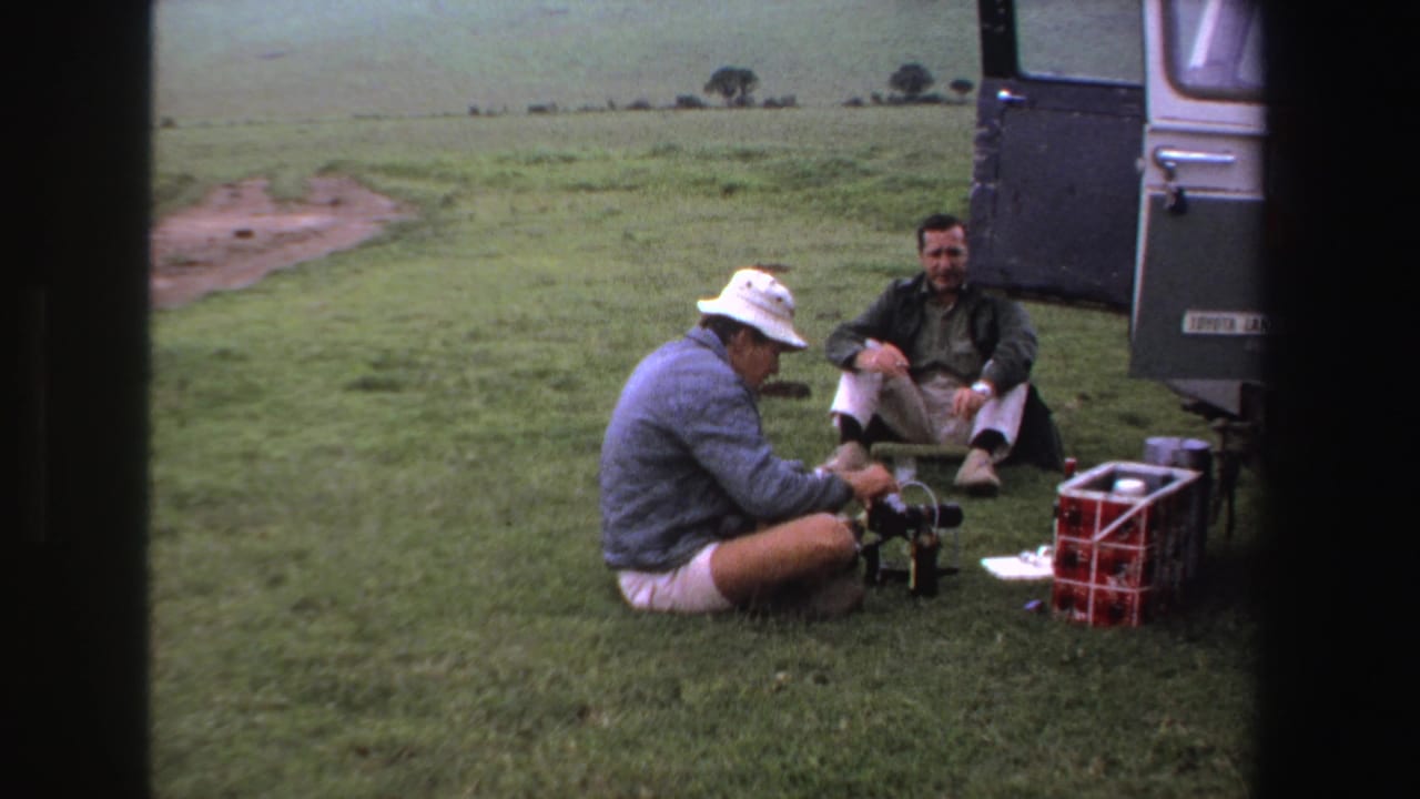 In 1969 South Africa footage, two adult men rest on grass beside an open utility vehicle as one adjusts a small camera setup.
