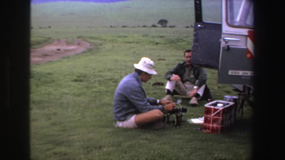 A man in a white hat adjusts camera equipment on a grassy plain while another man sits near the open vehicle door.