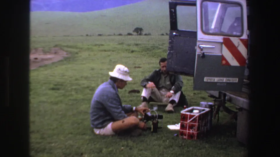 Two adult men are seated beside a Toyota Land Cruiser in a South African grassland setting, with a red crate and gear nearby.
