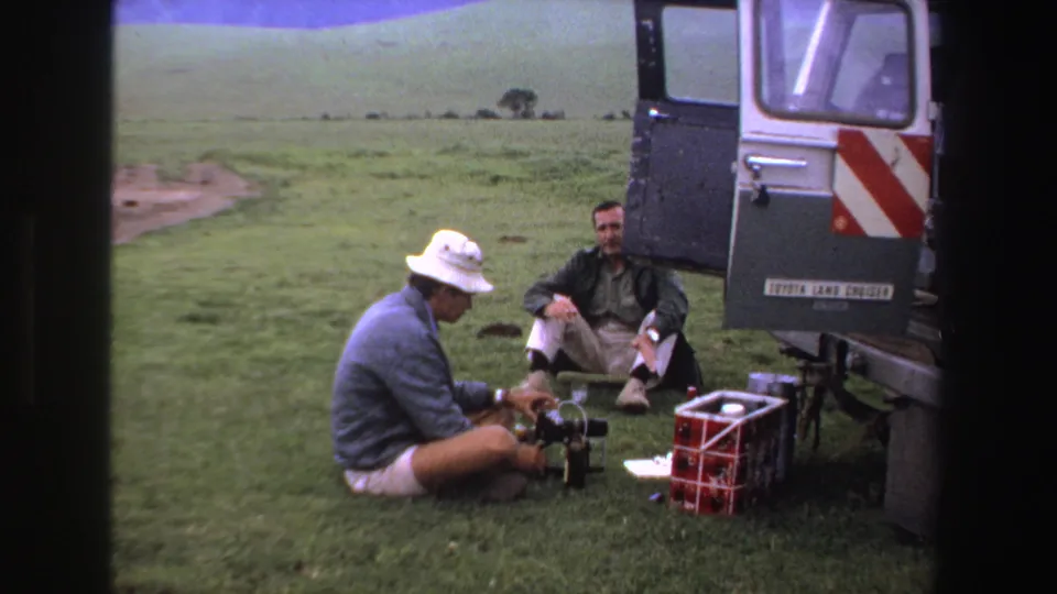 The frame shows two men beside an open utility vehicle on grass, with camera equipment and a red crate in the foreground.