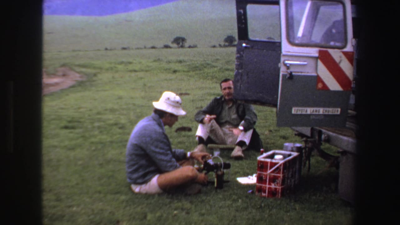 In 1969 South Africa, two adult men rest on a grassy plain beside a van with open doors, supplies, and camera equipment.