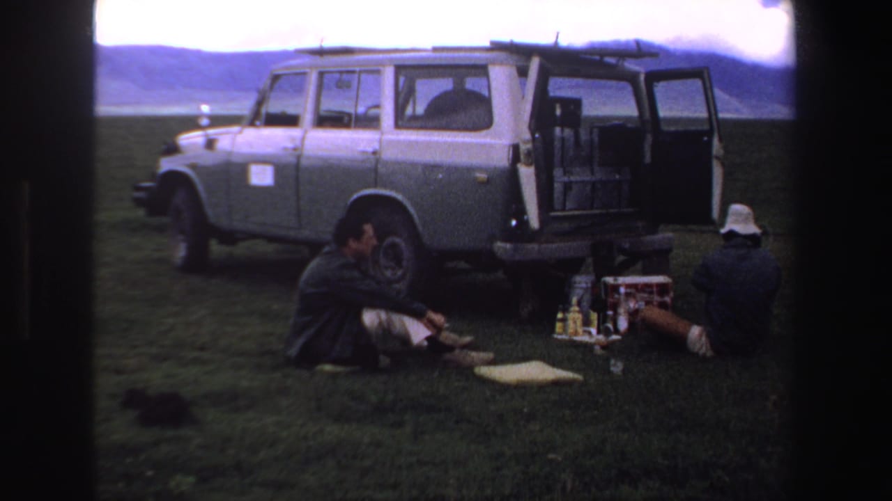 Grey van with rear door open as two men sit on grass with bottles and a cooler nearby.