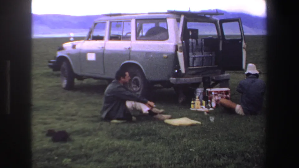 A 1969 South Africa travel frame shows two men seated beside a grey van, with picnic supplies on the grass and hills in the distance.