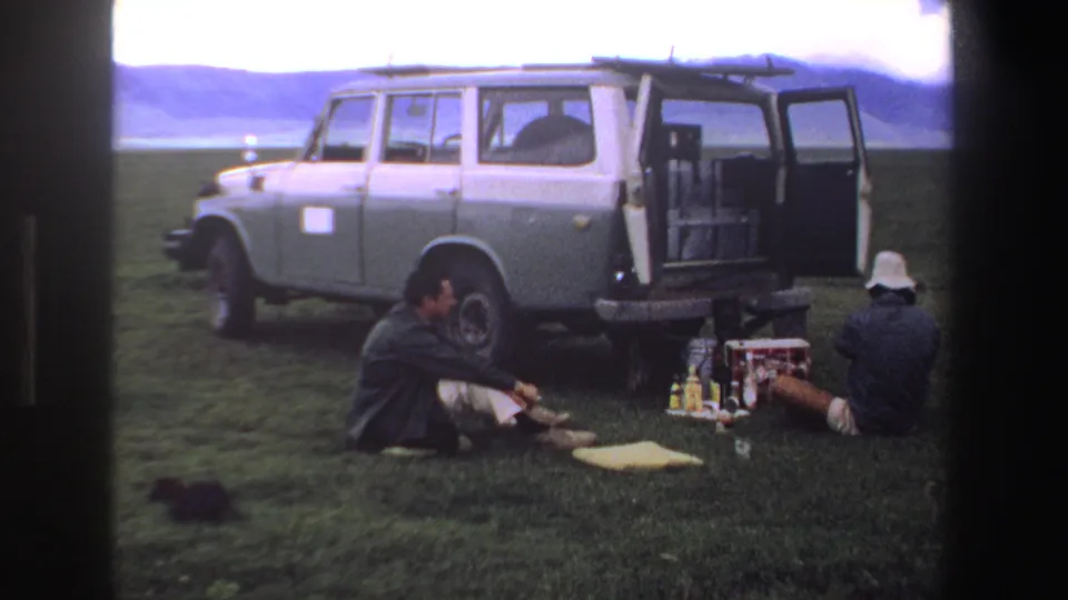 Two men rest beside a grey van in 1969 South Africa, with a cooler, bottles, grassland, and distant hills visible.