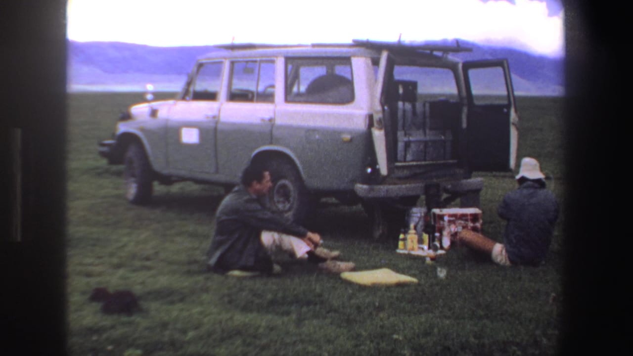 1969 South Africa home-movie frame showing two adults picnicking beside a grey station wagon on open grassland.