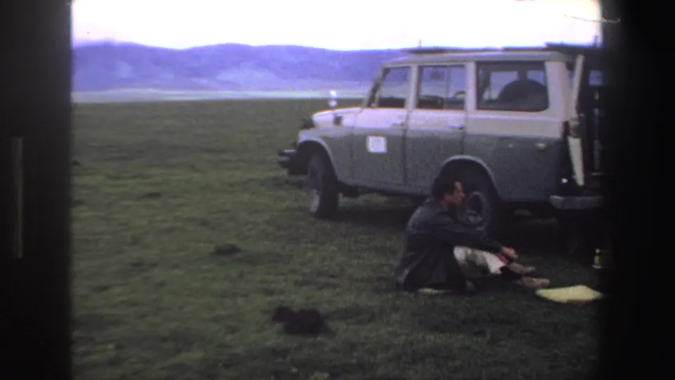 1969 South Africa frame with a man seated on grass beside a grey station wagon, with hills in the distance.