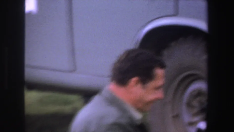 Adult man in a light jacket stands beside a vintage vehicle wheel in 1969 South Africa home-movie footage.