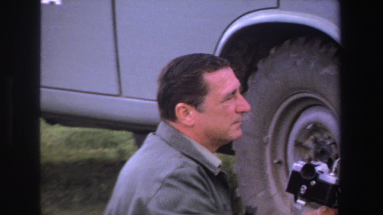 Man in profile holding a small camera near the wheel of a vintage vehicle.
