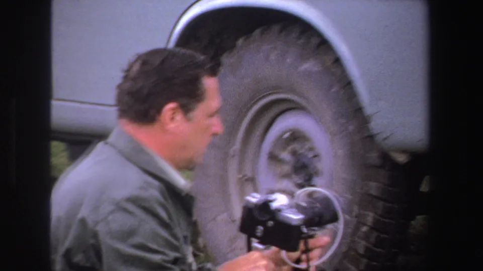 Adult man in a light jacket looks down at a camera beside the wheel of a vintage vehicle in 1969 South Africa footage.