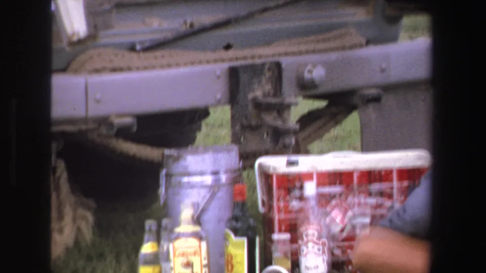 The rear area of the vintage vehicle appears with bottles, a cloth or basket, and repair items in the grass.