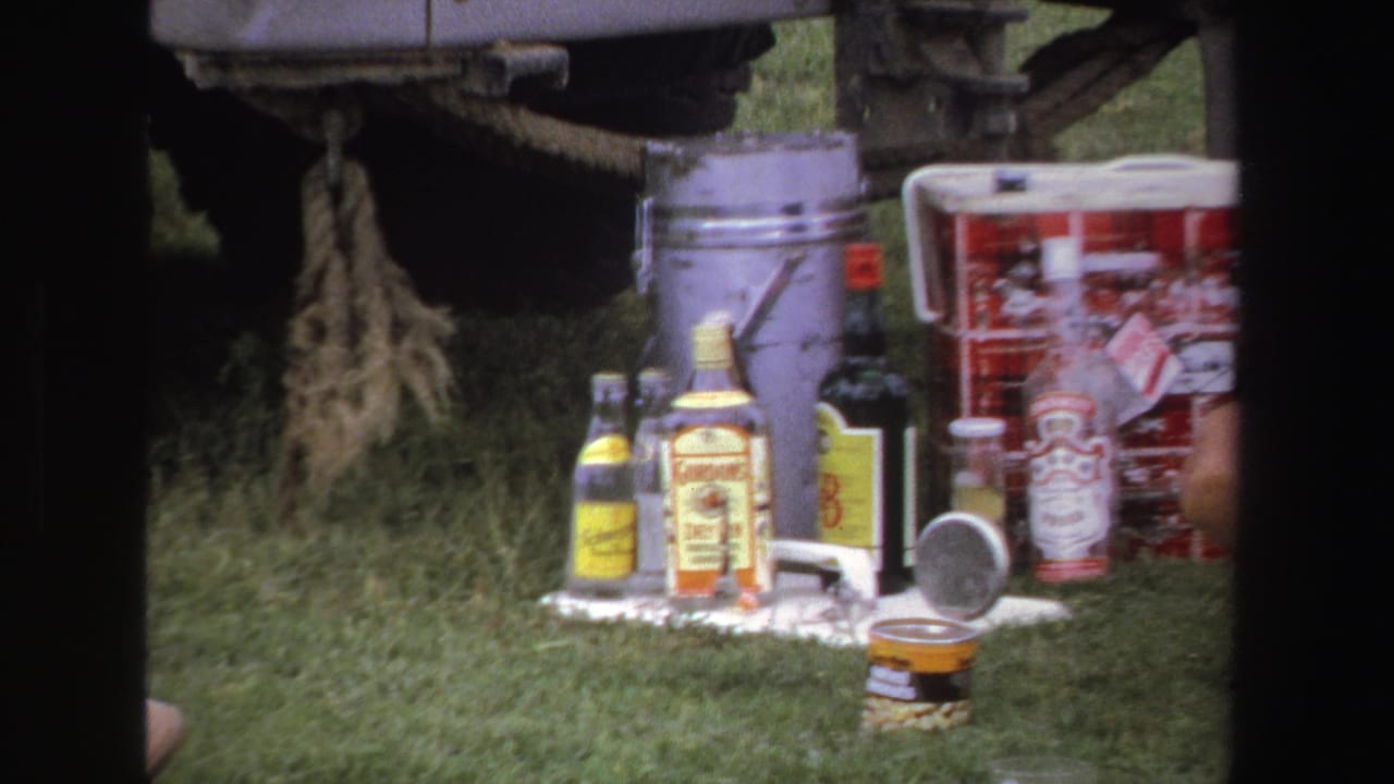 Picnic supplies, bottles, buckets, and red coolers set on grass beside a vehicle.