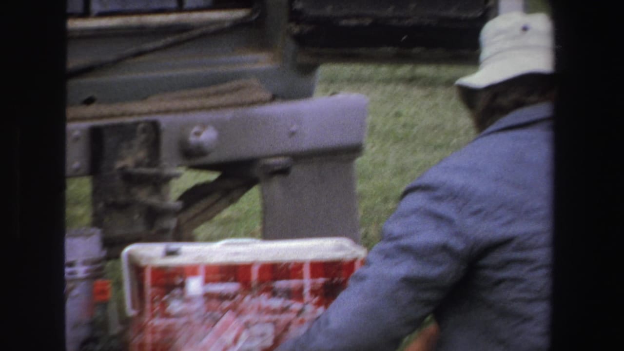 Adult in a white bucket hat sits near vehicle parts and red coolers on grass.