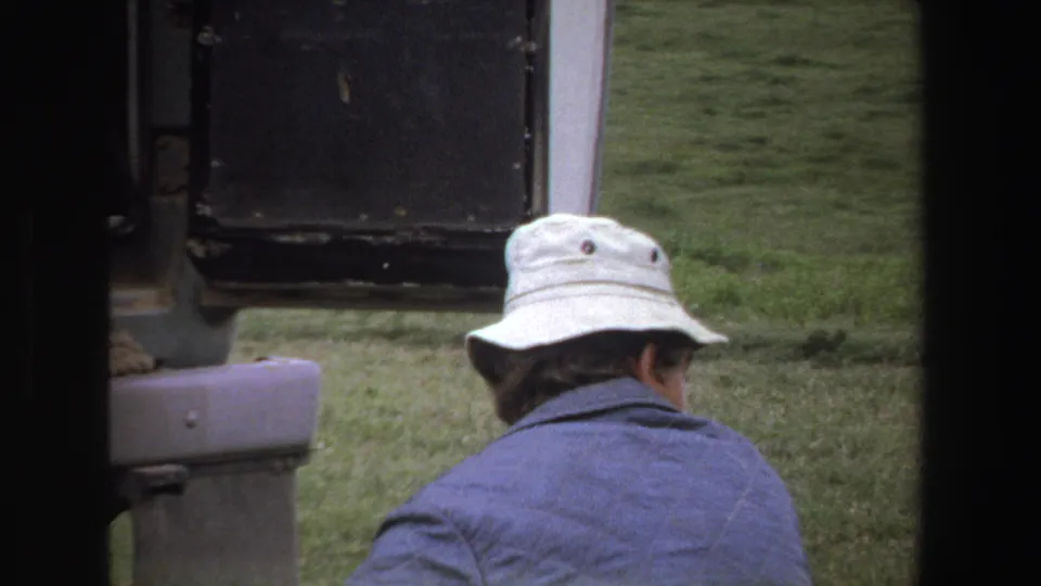 1969 South Africa 8mm frame showing an adult from behind in a white bucket hat and blue jacket near a vehicle on grass.