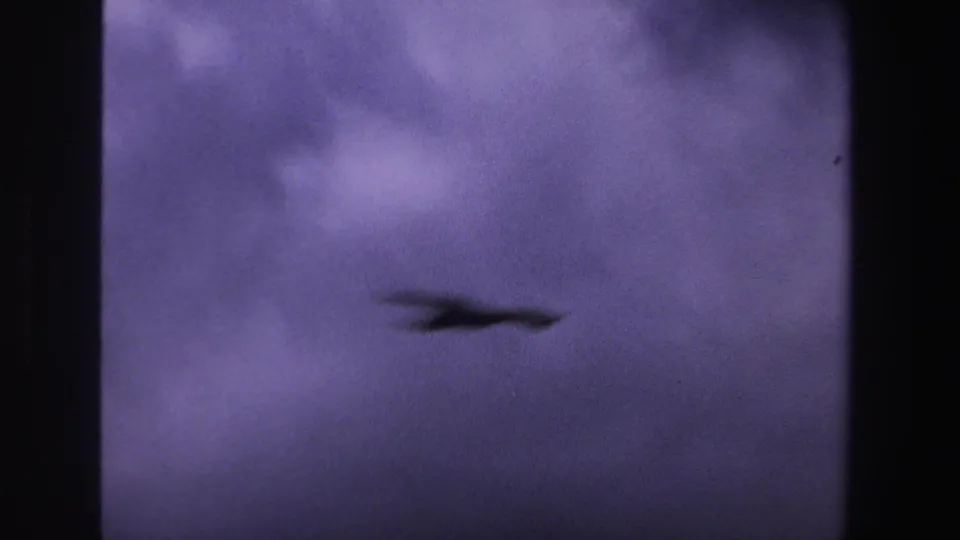 A blurred bird silhouette moves low across the frame beneath purple-tinted clouds in 1969 8mm footage.