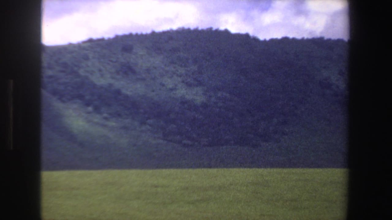 1969 South Africa home-movie frame with a green foreground field and dark hillside beneath an overcast sky.