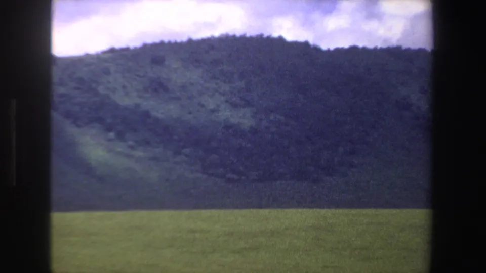 1969 South Africa home-movie frame with a green foreground field and dark hillside beneath an overcast sky.