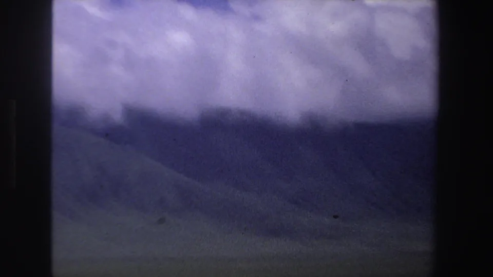 1969 South Africa 8mm frame showing low clouds, shadowed hills, and motion-softened grassland from a moving vehicle perspective.