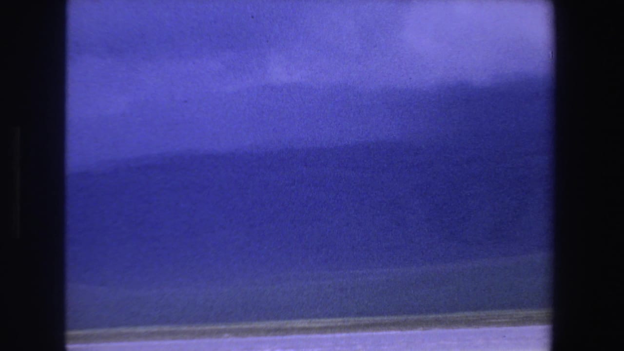 1969 South Africa 8mm frame with blue lake water, a distant mountain ridge, pale cloud cover, and shimmering light near the shore.