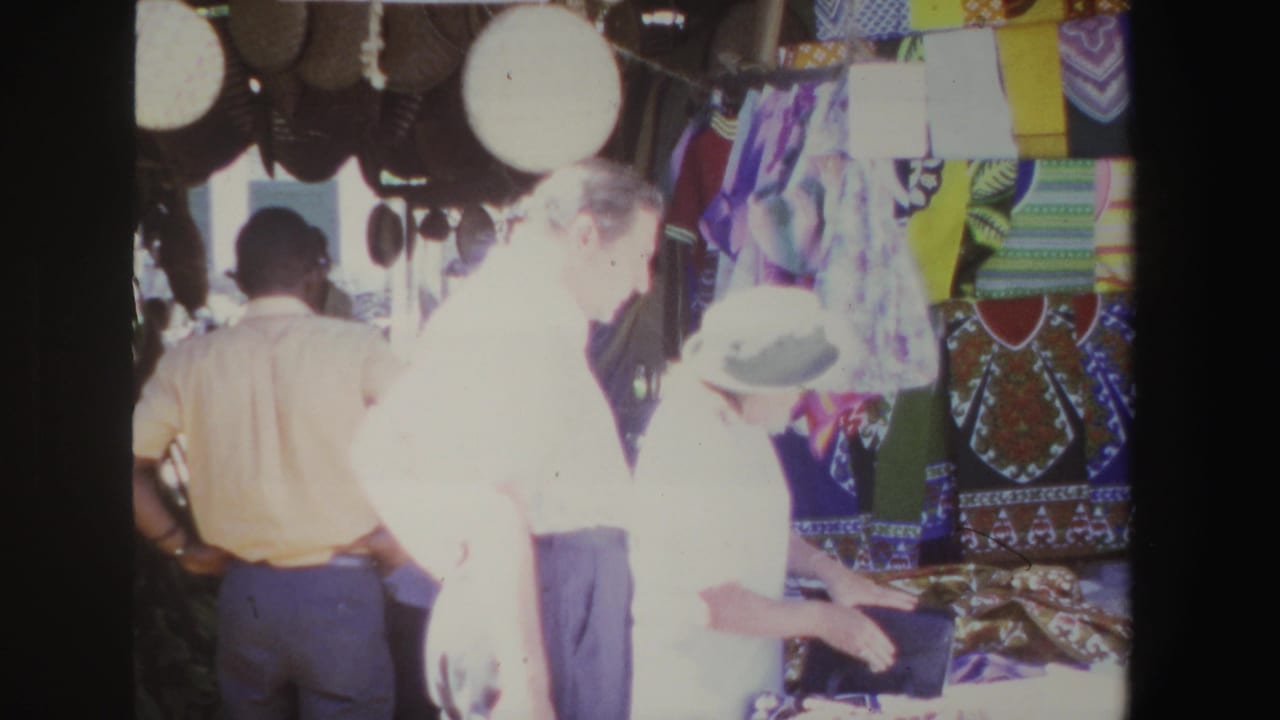 Adults browse colorful fabrics and hats at a shaded outdoor market stall.