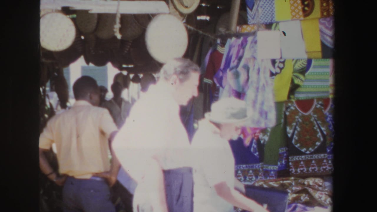 A woman in a light hat and sunglasses stands near a man by patterned textiles.