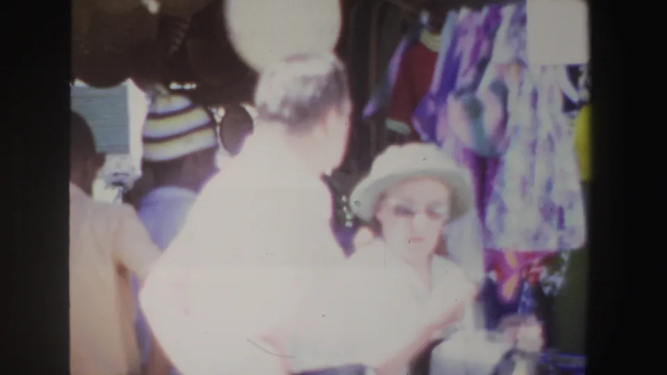 The camera moves closer as the woman in a hat and the man in a light shirt pass near hanging textiles and hats in the market.