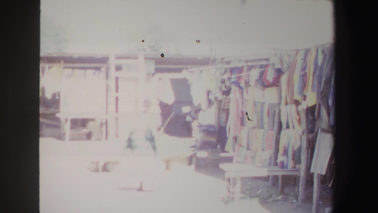 1969 South Africa home-movie frame showing a bright outdoor market stall with hanging clothing and a passerby in the foreground.