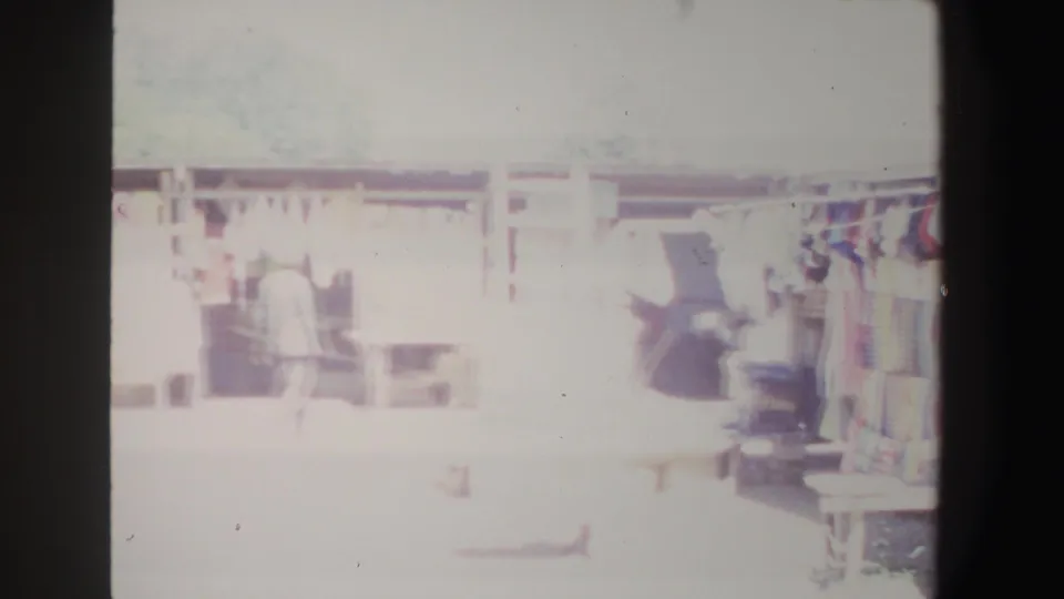 1969 South Africa market scene with a figure moving near shaded stalls, hanging textiles, and goods displayed along the walkway.