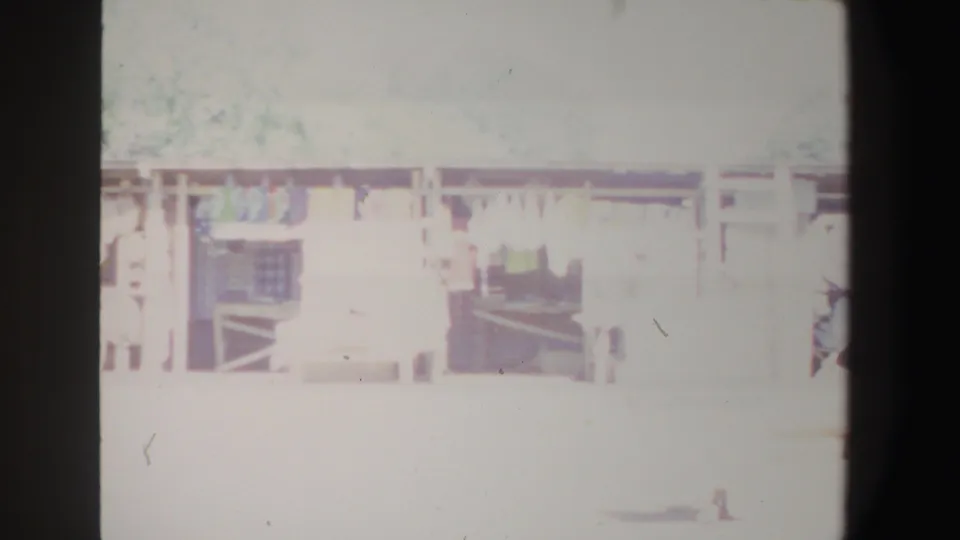 1969 South Africa home-movie frame of open-air market stalls with shaded shelving, goods, and indistinct figures in strong sunlight.