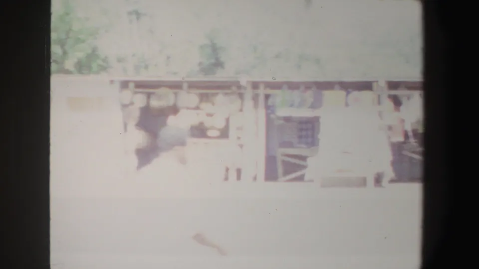 1969 South Africa market frame showing a person near stalls with round goods or pots and hanging items under a simple shelter.