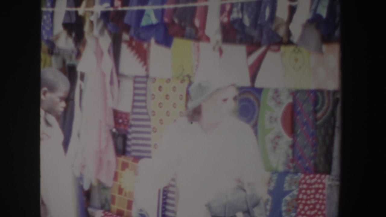 In South Africa 1969 home-movie footage, a woman in a white hat stands near colorful hanging textiles at a sunlit market stall.