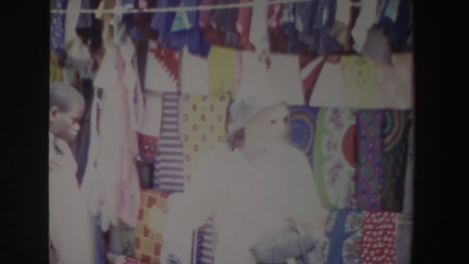 In South Africa 1969 home-movie footage, a woman in a white hat stands near colorful hanging textiles at a sunlit market stall.