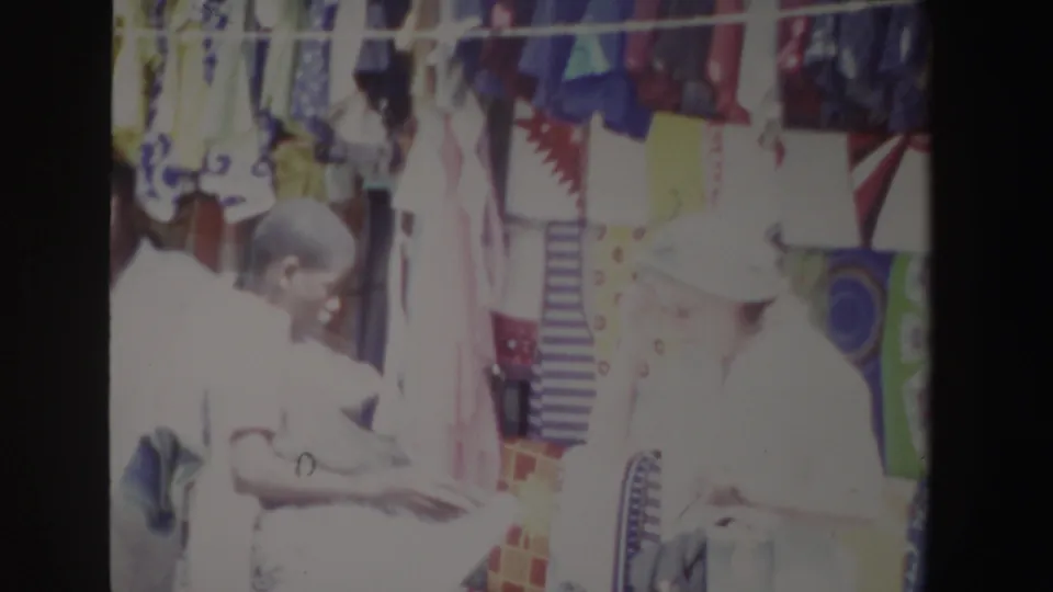 A sunlit South Africa 1969 market view shows a woman in white beside another person near dense rows of patterned fabrics.