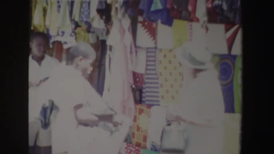 The 1969 South Africa market footage shows several adults near hanging fabrics as the woman in white appears to inspect merchandise.