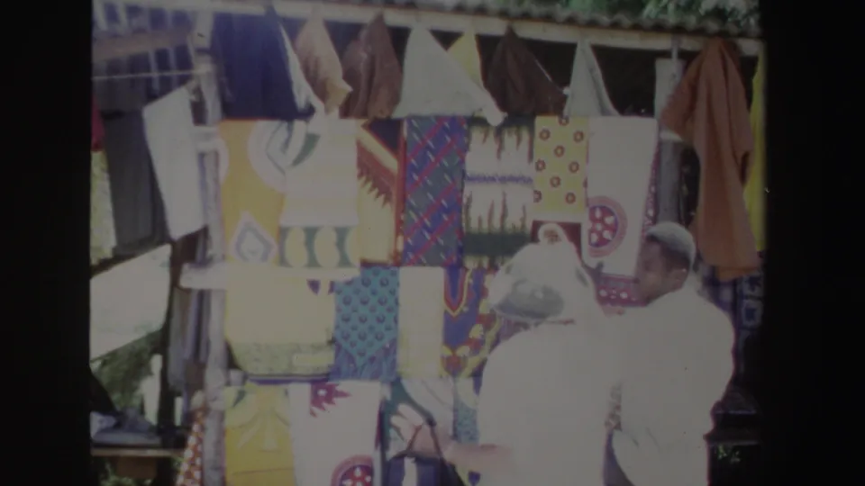 A wider 1969 South Africa market frame shows colorful textiles filling the stall while the woman in white stands at the edge of the display.