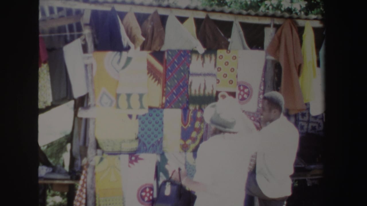 In South Africa in 1969, a woman in a light hat stands near a vendor at a bright outdoor textile display.
