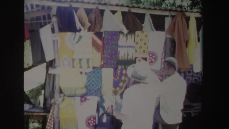 In South Africa in 1969, a woman in a light hat stands near a vendor at a bright outdoor textile display.