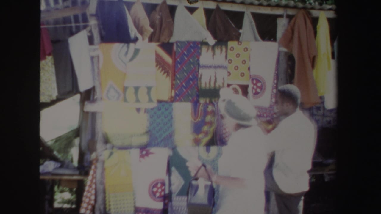 Vendor and woman stand close to a display of patterned cloths under a roof.