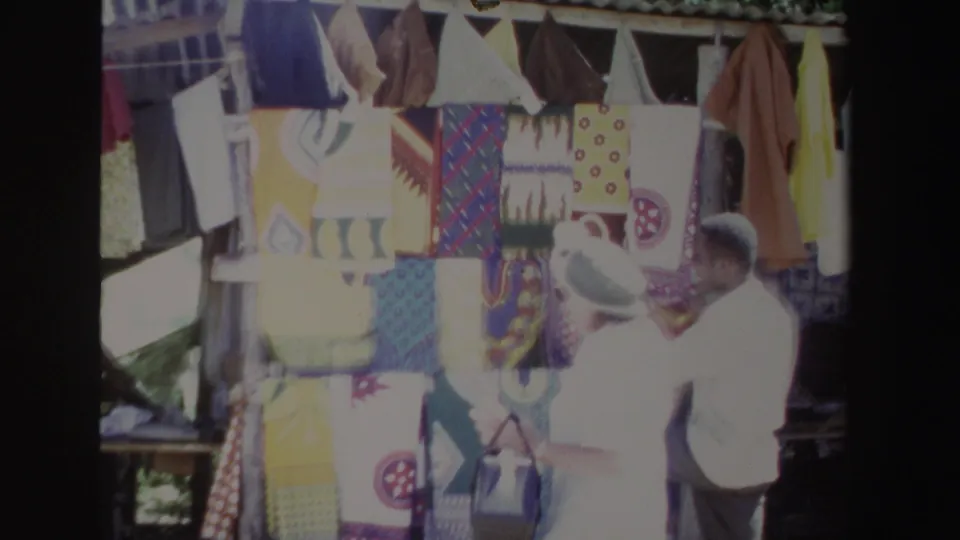 The woman and vendor look toward a colorful textile rack in a 1969 South Africa market scene.