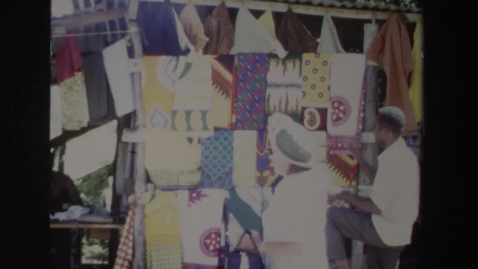A visitor in hat and shorts gestures toward hanging fabrics as the vendor watches at the South Africa stall.