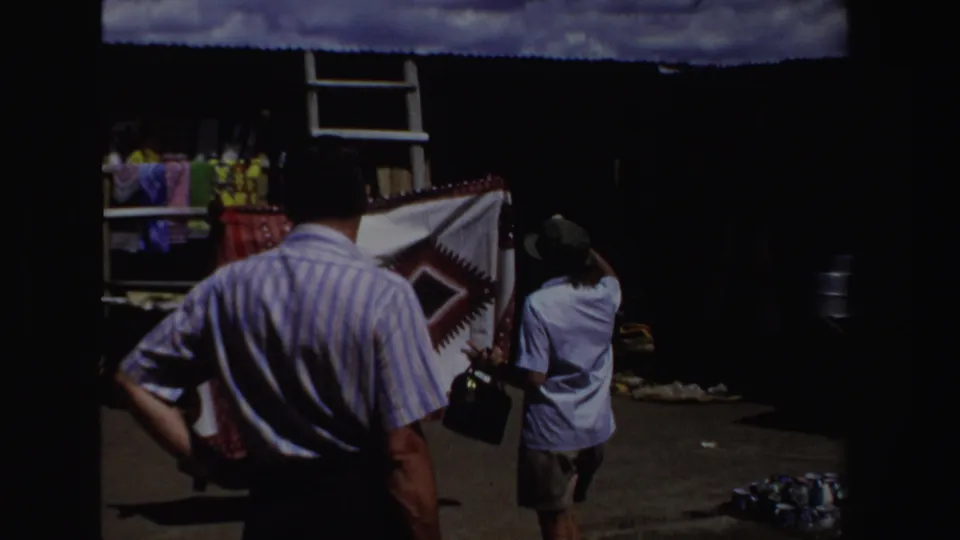 A wider South Africa market view shows two visitors or shoppers facing a shaded textile stall in 1969 footage.