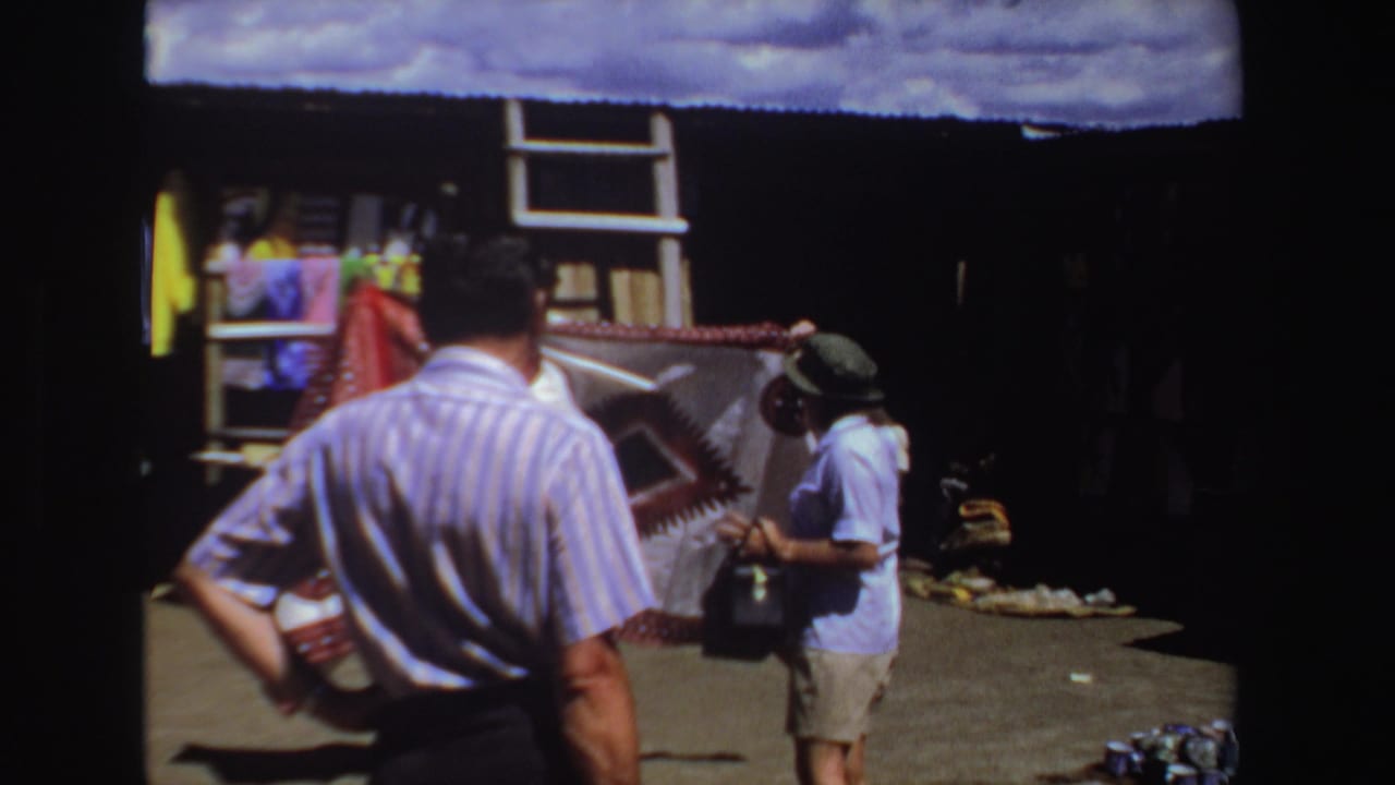1969 South Africa 8mm frame of tourists at an outdoor clothing stall as a seller holds up a patterned textile.