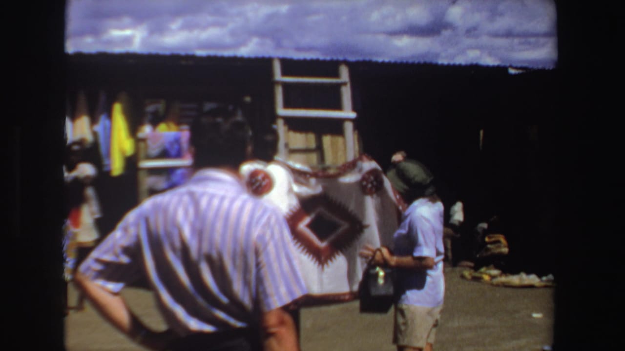 Tourists stand near a clothing stall while a seller displays a blanket or garment.