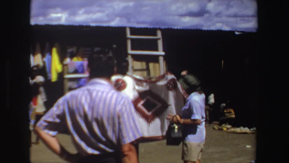 1969 South Africa market scene with a striped-shirt tourist and a seller displaying a patterned textile under a corrugated roof.