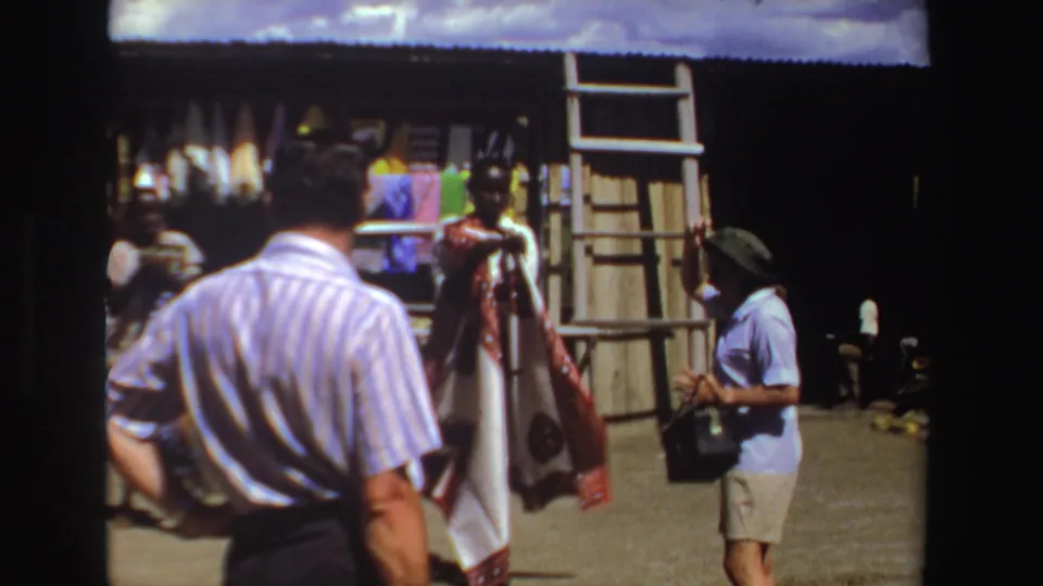 1969 South Africa 8mm frame showing a tourist in the foreground and a seller beside hanging clothing and textiles.