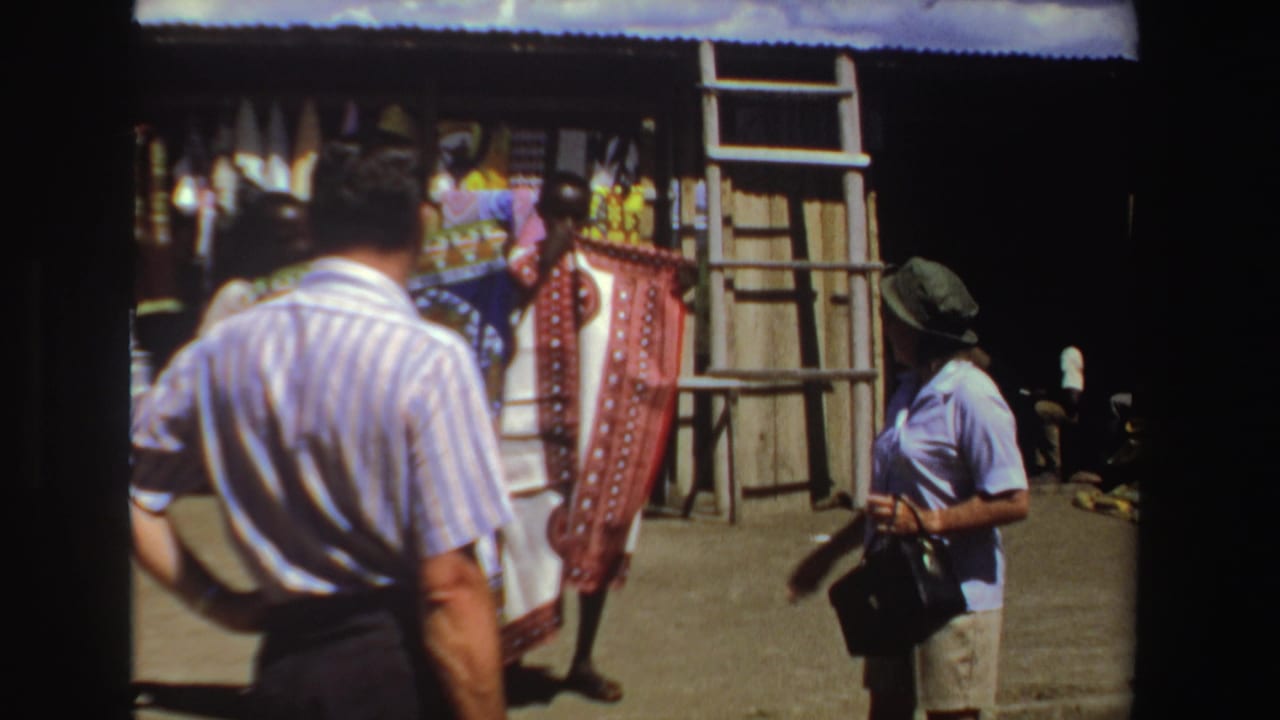 Tourists stand near a market stall with hanging textiles and a wooden frame.