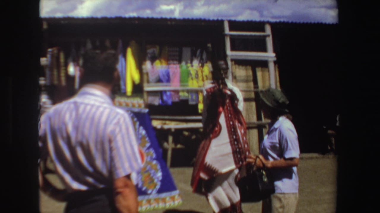 A man in a striped shirt faces a textile stall while another adult stands beside hanging cloth.