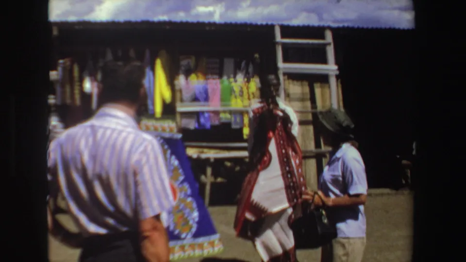 Tourist-shot 1969 South Africa market view with a striped-shirt man and colorful hanging fabric at a vendor stall.