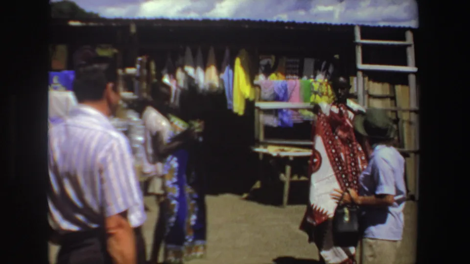 1969 South Africa home-movie frame of adults passing a market stall with colorful cloth and craft displays.